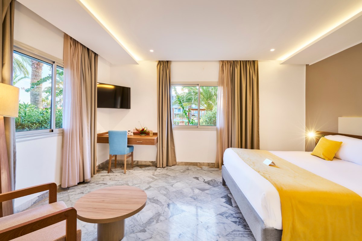 Modernes Hotelzimmer mit hellen Wänden, großem Bett, blauem Sessel und elegantem Beleuchtungskonzept.