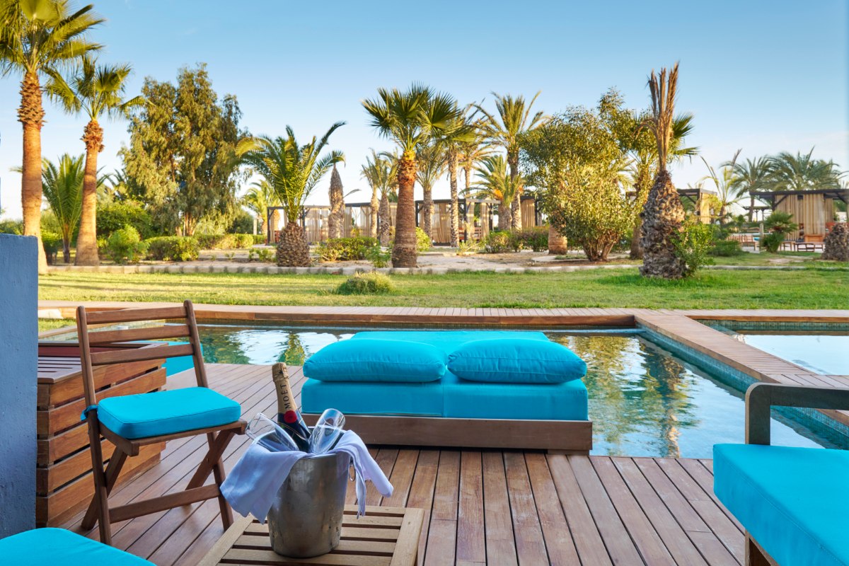 Modernes Hotel Wohnzimmer mit bequemer grauer Couch, rundem Esstisch und blauen Stühlen, hellem Marmorboden und minimalistischer Einrichtung.