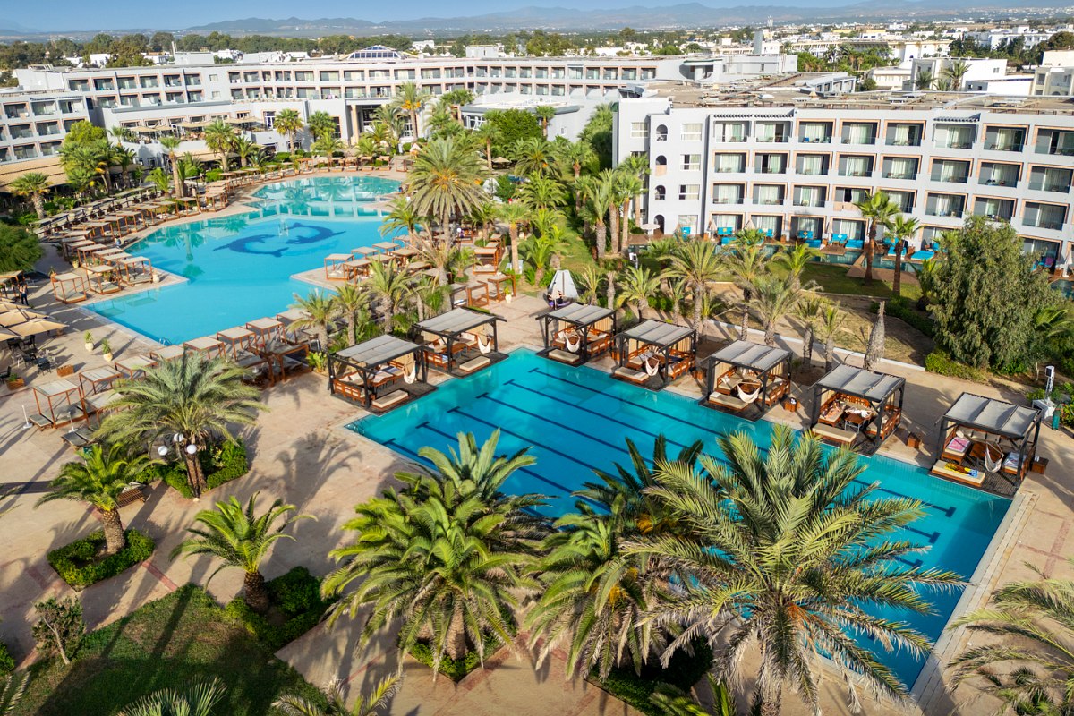 Sonnige Außenlounge im Hotel mit überdachten Holzpavillons, gemütlichen Sitzgelegenheiten und palmenumgebenem Ambiente.