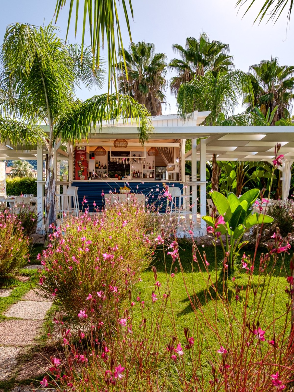 Garten mit pinken Blumen, viel Grün, Palmen und Bistro-Bar mit blauen Thekenfront und weißen Möbeln.