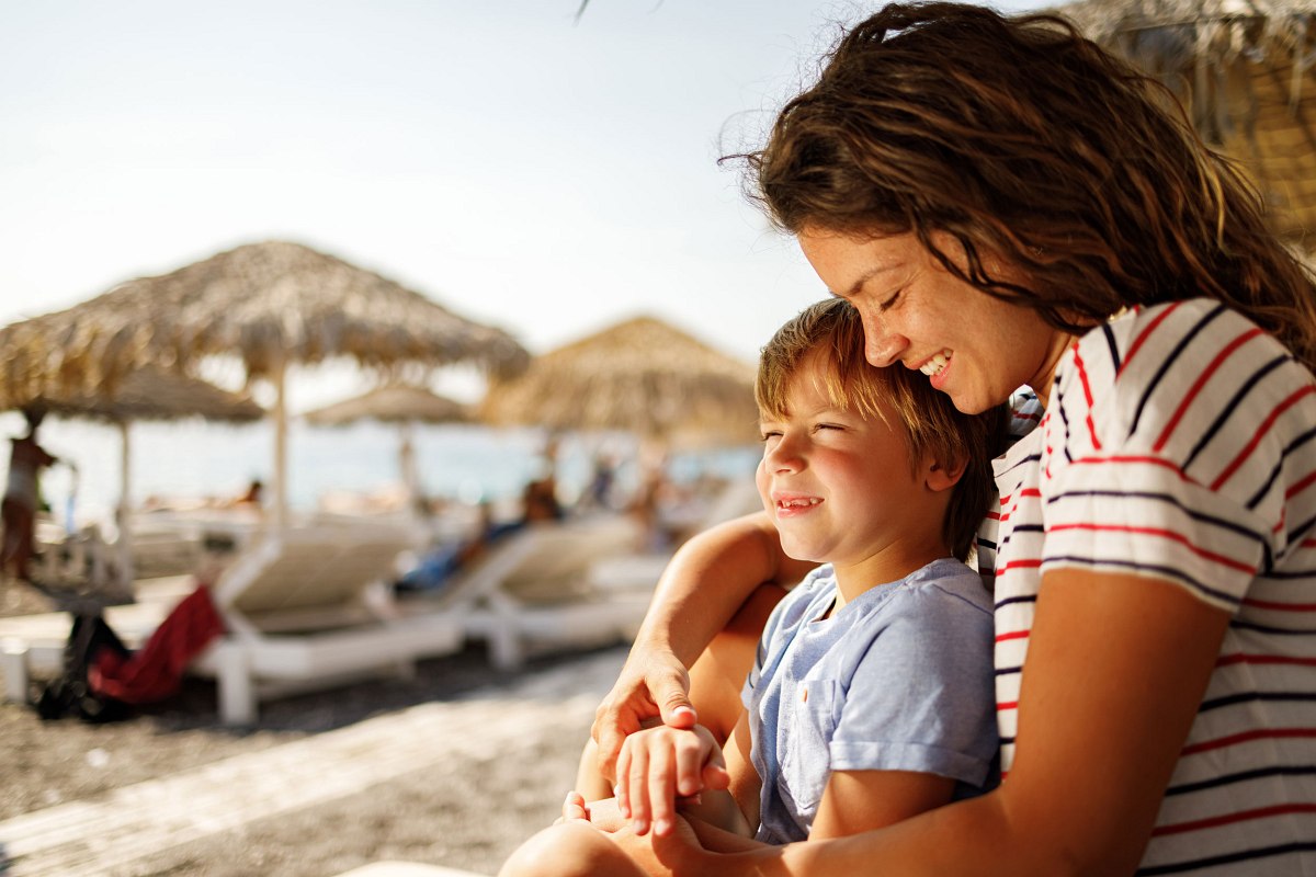 Mutter und Kind genießen lachend die Sonne am Strand – inniger Moment unter Strohdächern und vor Meereskulisse.