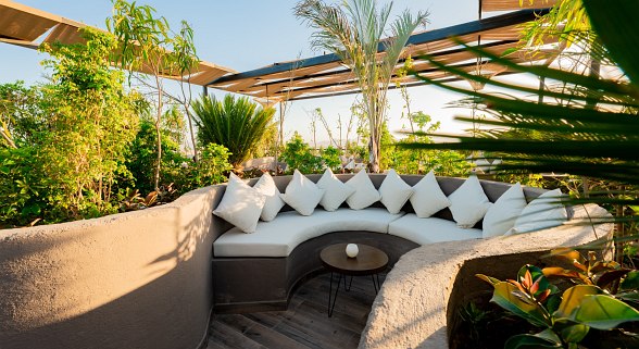 „Gemütliche Lounge-Ecke mit weißen Kissen, umgeben von tropischem Grün und überdacht von Holzlamellen.“