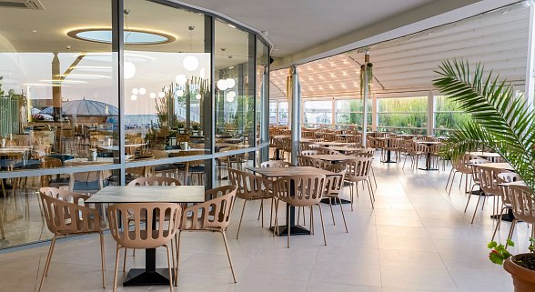 Restaurant mit Außenbereich: geräumiges Restaurant mit großen Fenstern, Tische und Stühle zur Außenterrasse, überdachte Außenbereich, gemütliche Atmosphäre mit Pflanzen und Meerblick.