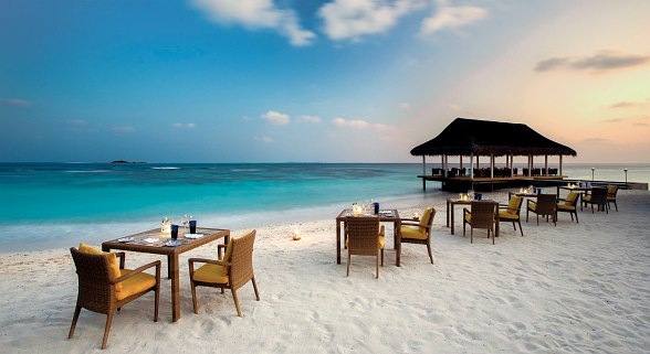Tische und Stühle mit gemütlichen Polstern am weißem Sandstrand, direkt am türkisfarbenen Meer, mit kleinem, überdachtem Strandrestaurant im Hintergrund.