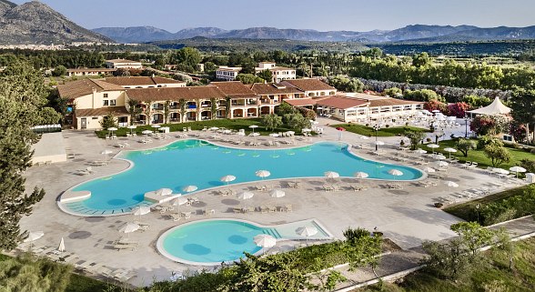 Gemütliche Hotelanlage mit großer Poollandschaft, Sonnenschirmen, Liegen und mediterranen Gebäuden in grüner Umgebung und hügeligem Bergpanorama im Hintergrund.