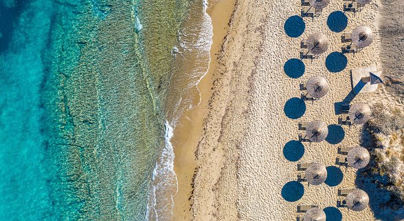Drohnenaufnahme eines Sandstrands mit türkisblauem Meer, Sonnenschirmen und klaren Schatten bei hellem Sonnenlicht.
