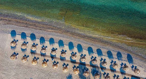 Draufsicht auf Strand mit gleichmäßig angeordneten Strohdach-Sonnenschirmen und Liegen am türkisfarbenen Meer.