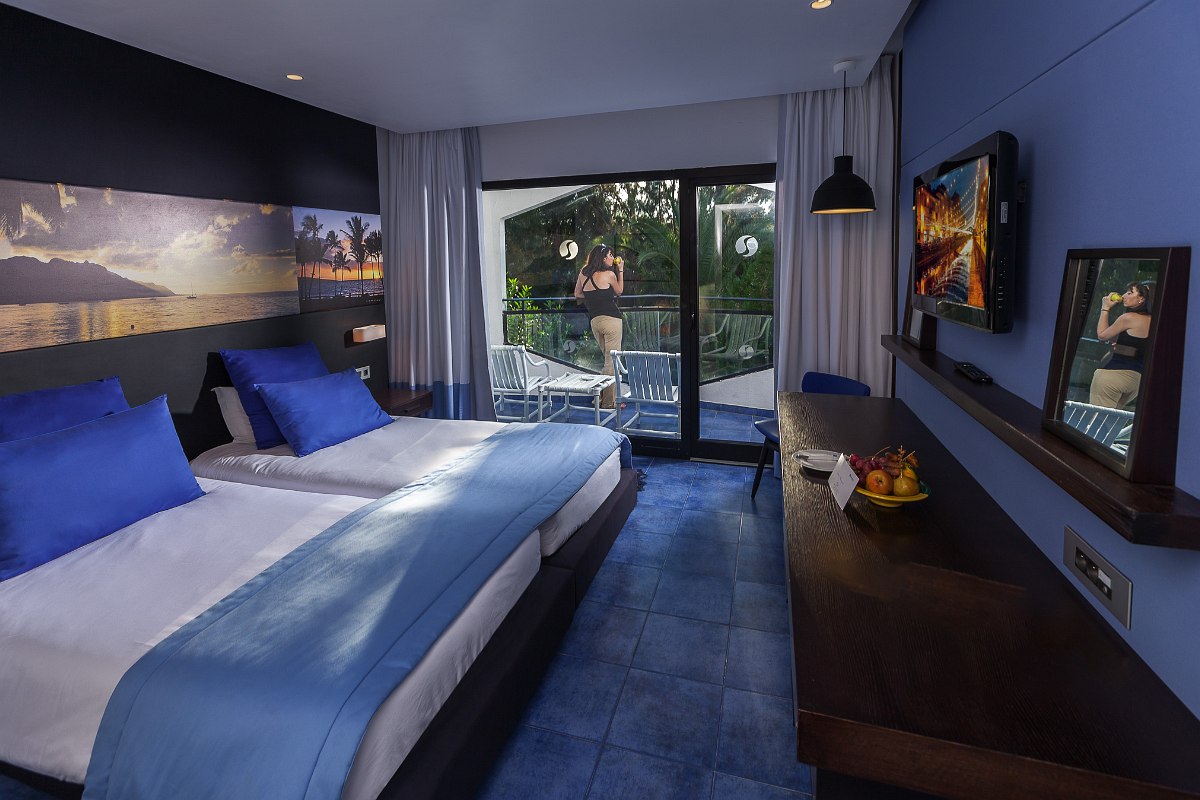 Hotelzimmer mit zwei Einzelbetten, blauen Bettdecken und Kissen, blauen Fliesenboden, Fernseher an der Wand und Balkon mit Blick ins Grüne.