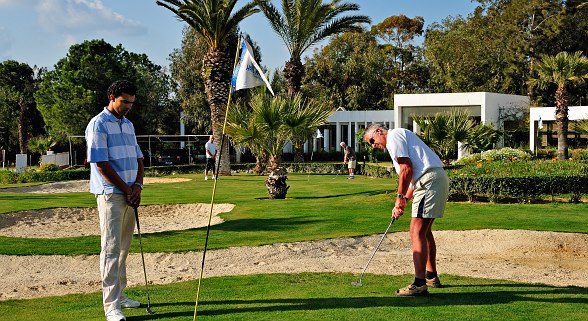 Vier Männer spielen Golf auf einem gepflegten Golfplatz mit Palmen und modernen Gebäuden im Hintergrund.