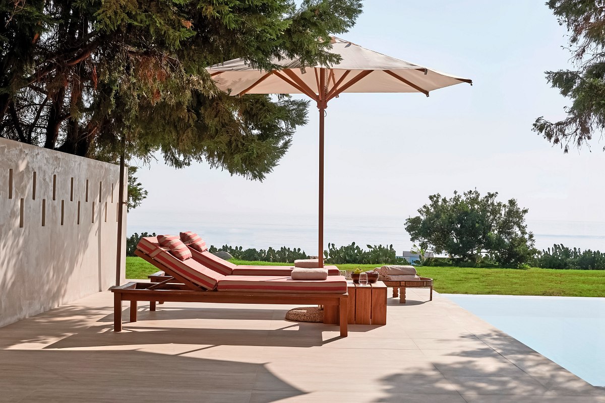 Elegante Sonnenliegen mit Schirm auf einer Terrasse am Pool, umgeben von Grün und mit Blick auf das ruhige Meer.
