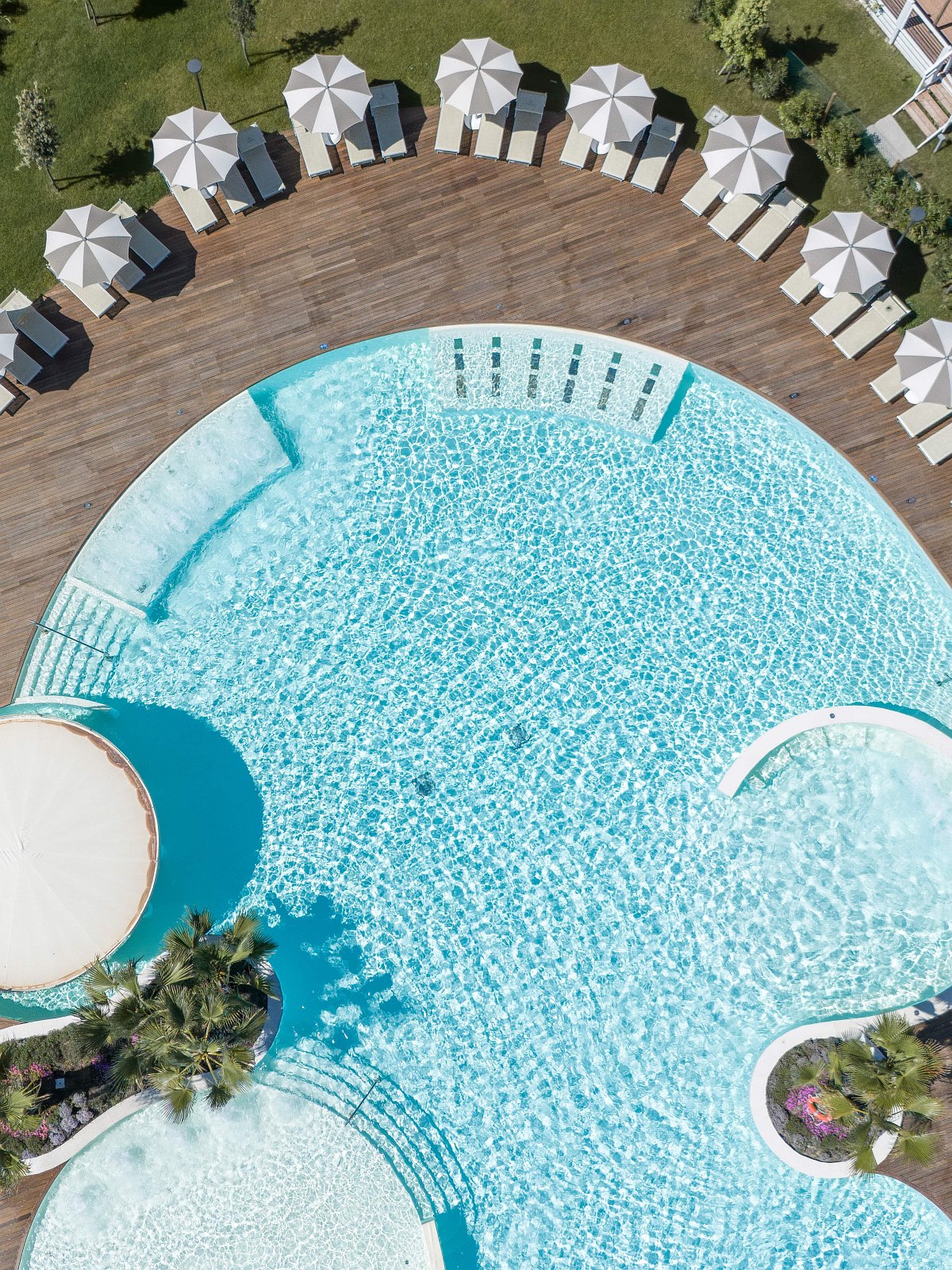 Drohnenaufnahme eines großen, runden Pools mit Sonnenliegen und Schirmen auf Holzdeck, umgeben von Palmen und grüner Anlage.