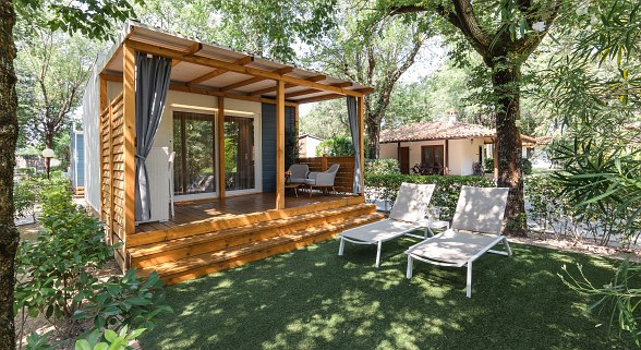 Gemütliches Mobilheim mit überdachter Holzterrasse, Sitzgruppe, Liegestühlen und schattigem Garten – naturnah und einladend.