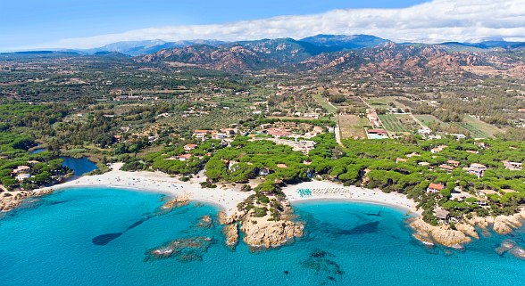Hotelanlage an zwei Buchten mit weißem Sandstrand und kristallklarem Wasser in grüner Umgebung und hügeligem Bergpanorama im Hintergrund.