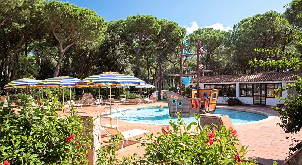 Kleiner Pool mit Piratenschiff, umgeben von Sträuchern und großen Bäumen. Liegen und Sonnenschirme stehen um den Pool herum, im Hintergrund befindet sich ein kleines Gebäude.