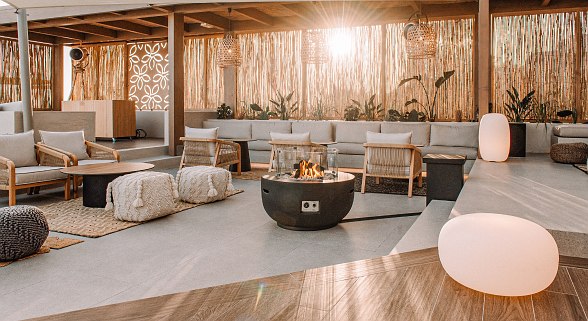 Lounge mit gemütlichen Sofas und Sesseln, Feuerstelle in der Mitte, natürlicher Deko und Sonnenlicht im Hintergrund.