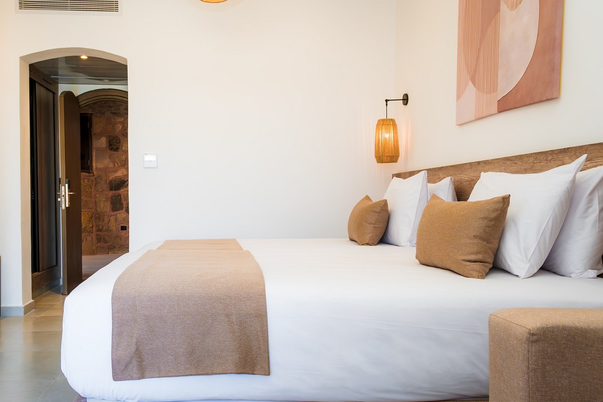 Stilvolles Hotelzimmer mit Doppelbett, blauen Akzenten, gewölbter Backsteindecke und Balkon mit Blick auf eine Resortanlage.