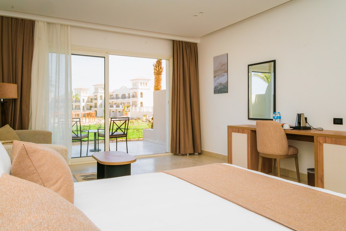 Stilvolles Hotelzimmer mit Doppelbett, blauen Akzenten, gewölbter Backsteindecke und Balkon mit Blick auf eine Resortanlage.