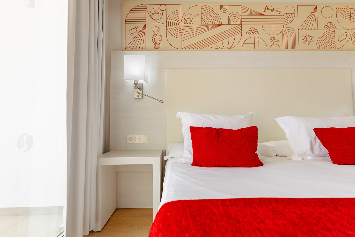Bright hotel room with double bed, red pillows, flat-screen TV, desk and balcony with view of palm trees and building.