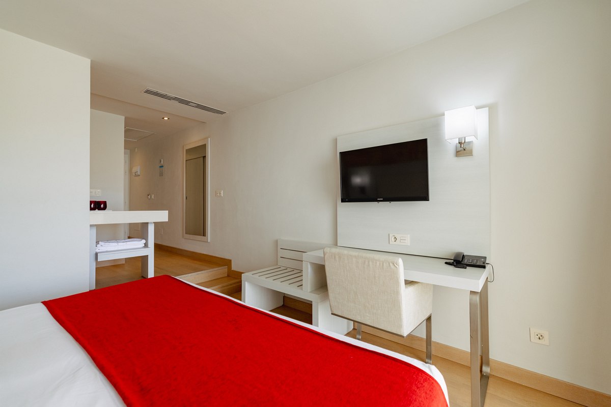Modern hotel room with open bathroom, large shower, red mosaic mirror, double bed, TV and balcony with seating area.