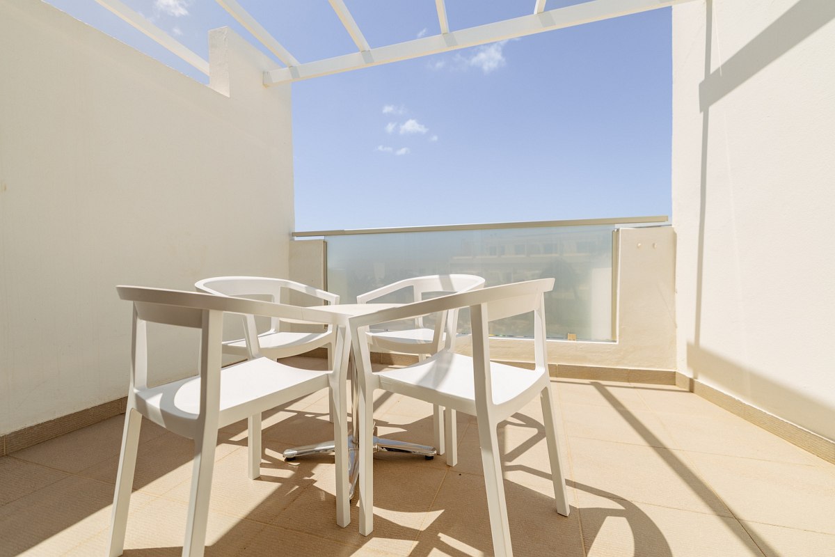 Bright hotel balcony with two white chairs and table, sea view and modern white pergola under a blue sky.