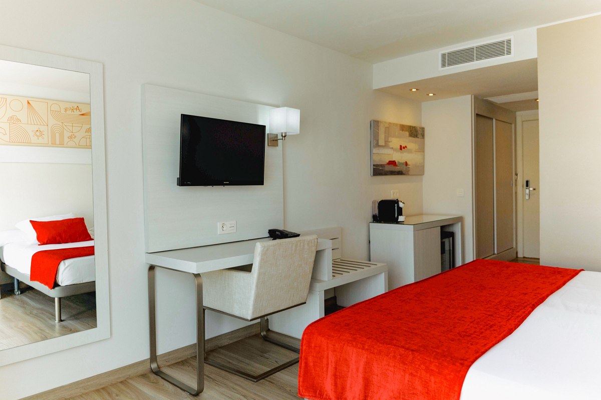Bright hotel room area with desk, TV, coffee machine, kettle, minibar and curtains in front of the window.