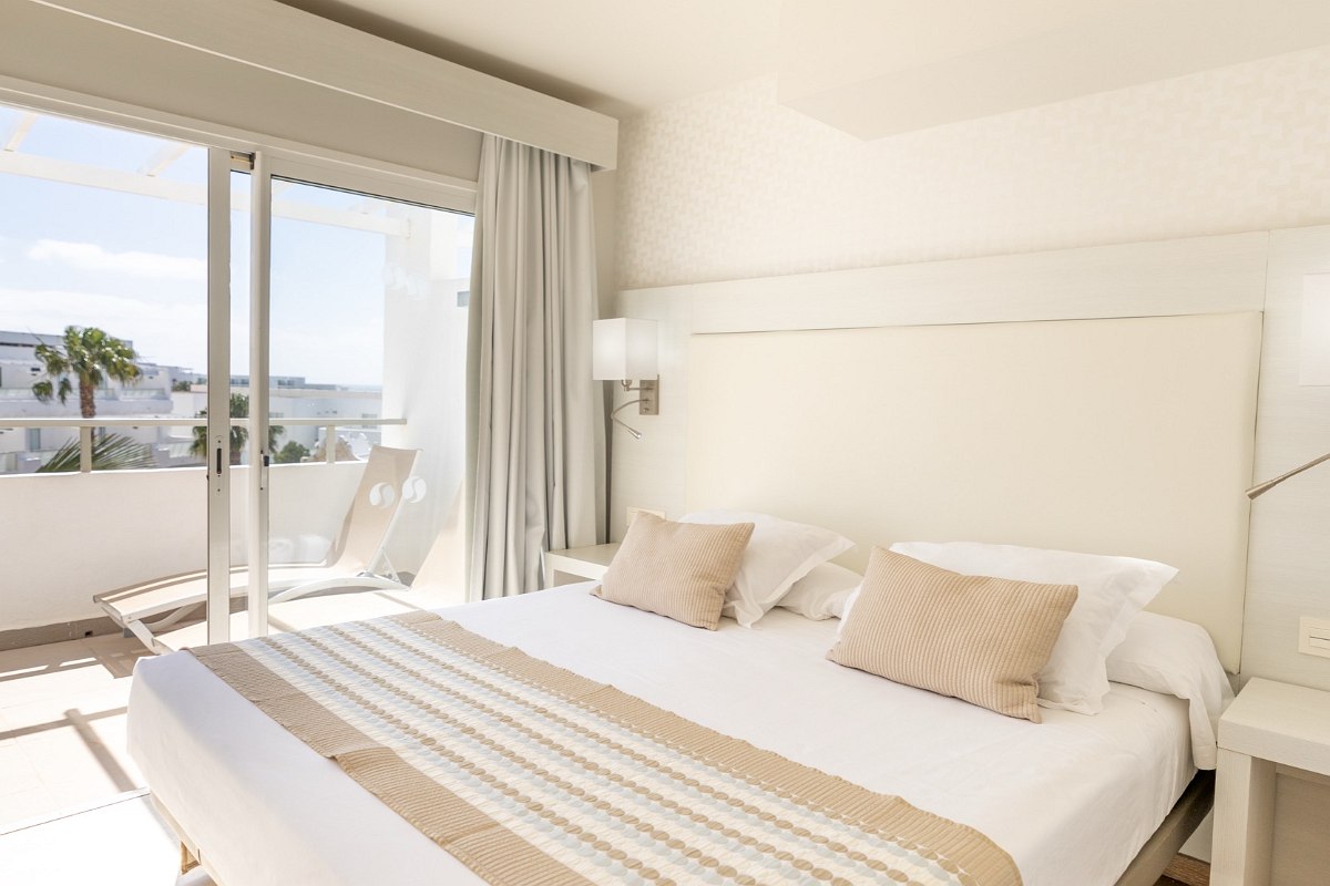 Bright hotel room with double bed, turquoise pillows, balcony with sun loungers and view of white buildings and blue sky.