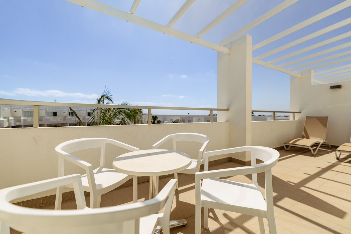 Modern hotel balcony with white table and chairs, two loungers, glass railings and a view of the blue sky.