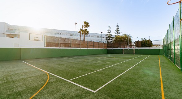 Multifunctional sports field with green flooring, goals, basketball hoop and tennis net in front of a white hotel building in the sunshine.