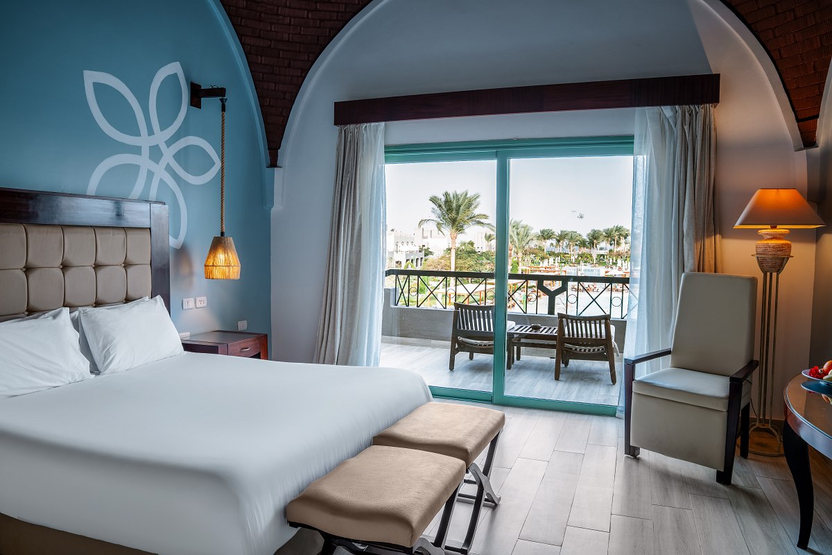 Spacious room with vaulted brick ceilings, large bed with red accents, balcony overlooking a scenic view, and elegant decor.