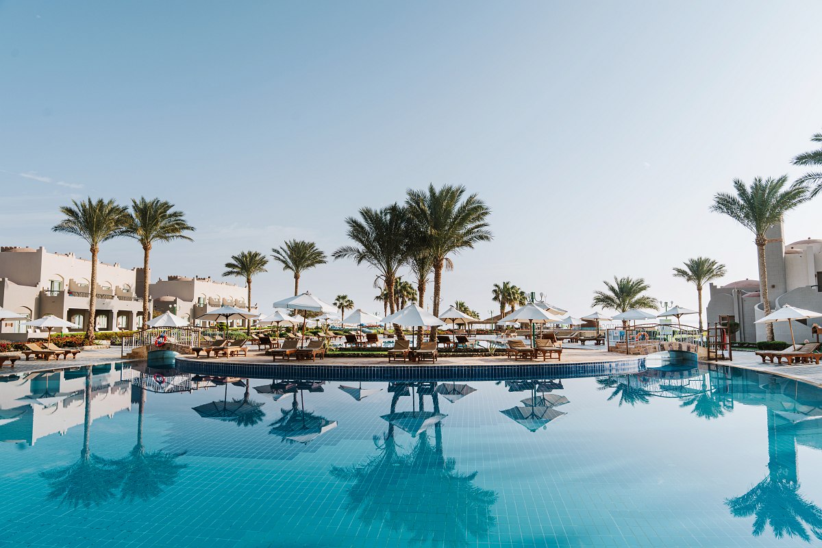 a hotel complex with castle-like architecture, illuminated pool, palm trees and atmospheric reflection in the water.