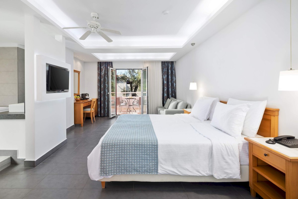 Cozy bedroom with a wooden double bed featuring white linens and a yellow throw. A nightstand holds a phone and mug. Sliding glass doors open to a patio with a table and two chairs, overlooking a lush green lawn.