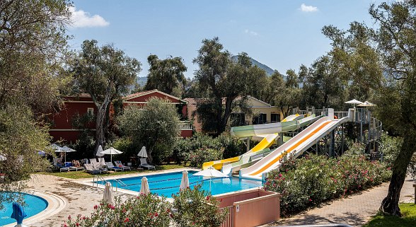 Outdoor water park with colorful slides, a pool, surrounded by trees and flowers. Comfortable lounge chairs and umbrellas provide relaxation spots.