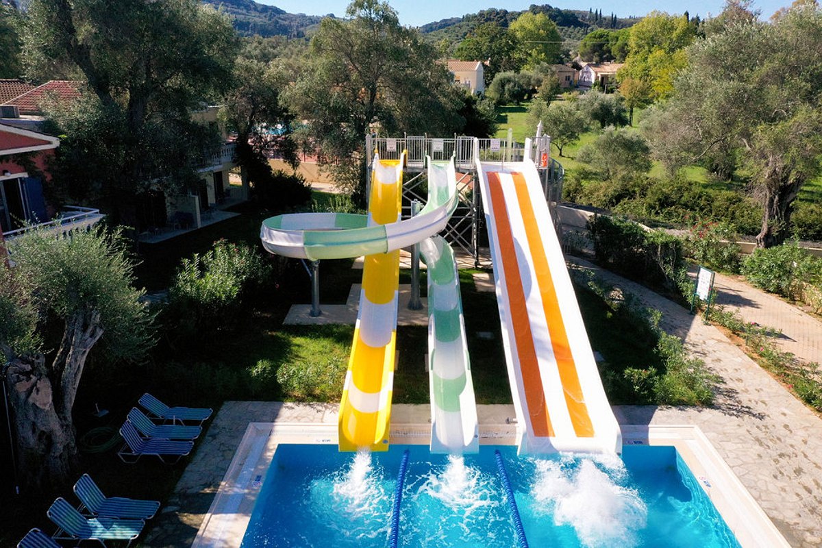 Evening view of a large outdoor pool surrounded by dining and bar areas, warmly lit with ambient lights and a wooden bridge crossing part of the pool.