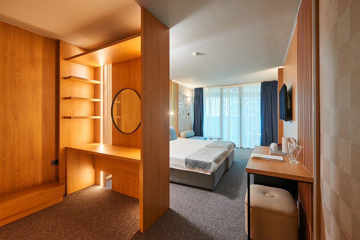  Sentido hotel room with walk-in wooden closet, integrated mirror and shelves, bed, desk and balcony with blue curtains.