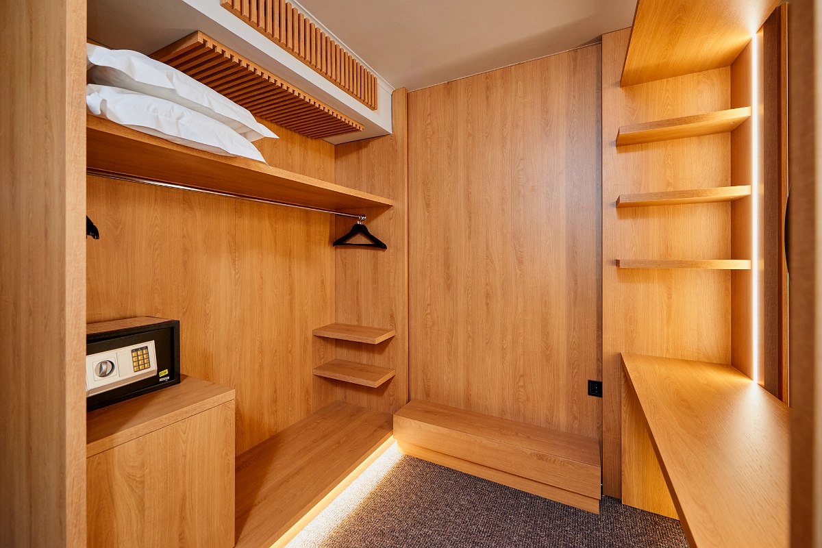 Walk-in wooden closet with safe, shelves, clothes hangers and bench in warm light.