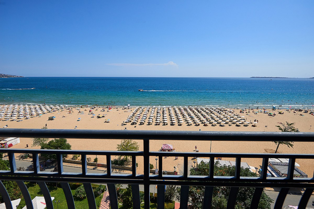 Blick vom Balkon auf Sandstrand mit Sonnenschirmen, Liegen und blauem Meer unter klarem Himmel.