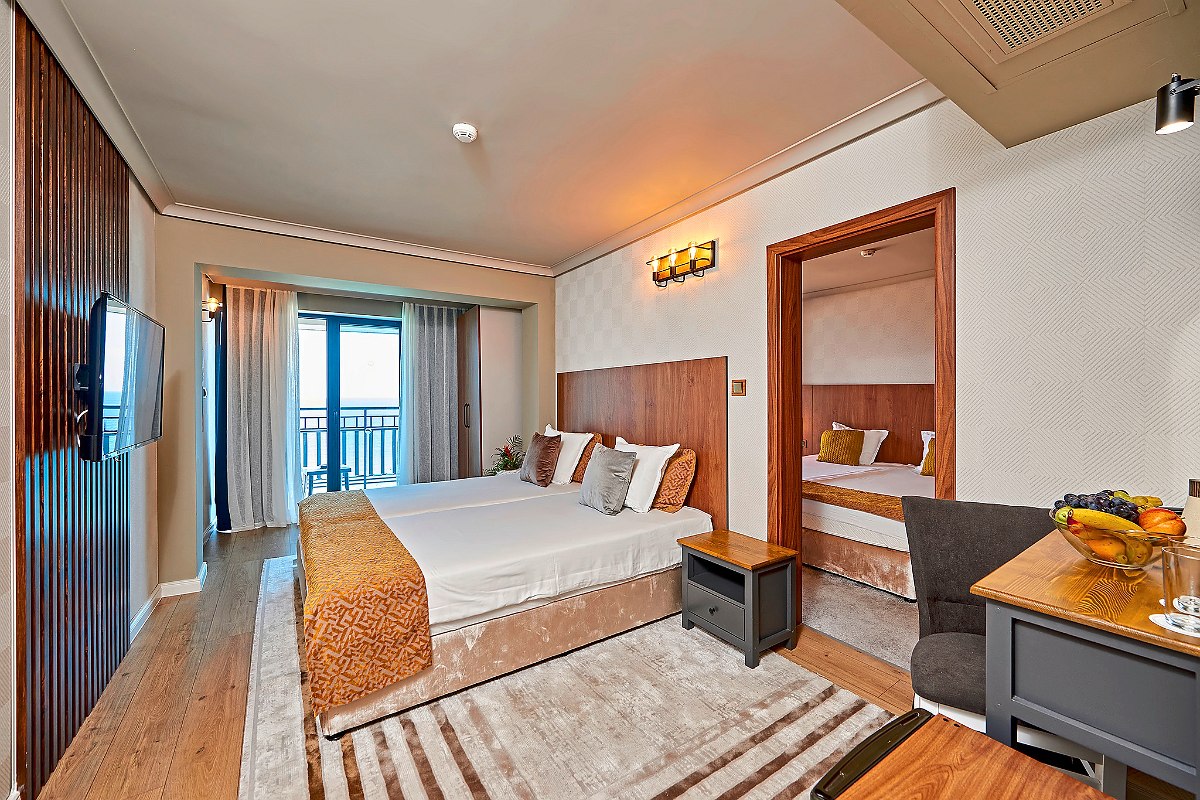 Family room Sentido Bellevue Beach: Modern hotel room with double bed, connecting door to separate bedroom, flat-screen TV, balcony and desk with fruit basket.