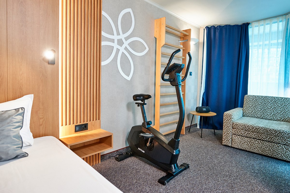 Close-up of the room with bed, bedside table, wall bars, fitness bike, couch, blue curtain, Bluetooth music box and small side table.