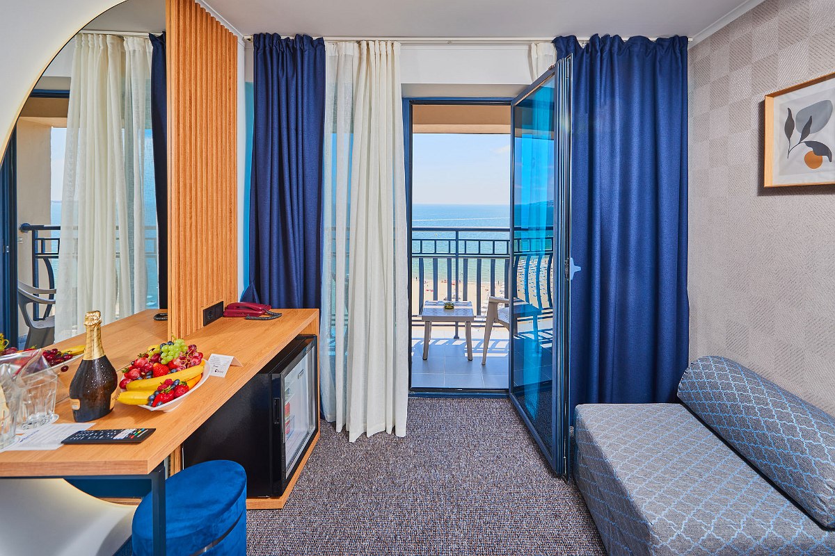  Double hotel room with seating area, balcony with sea view, table with champagne and fruit, blue curtains.