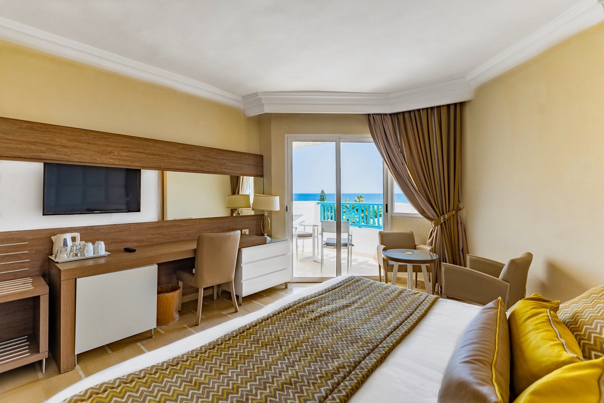 Bright hotel room with double bed, balcony with sea view, desk, seating area and modern wooden furniture.