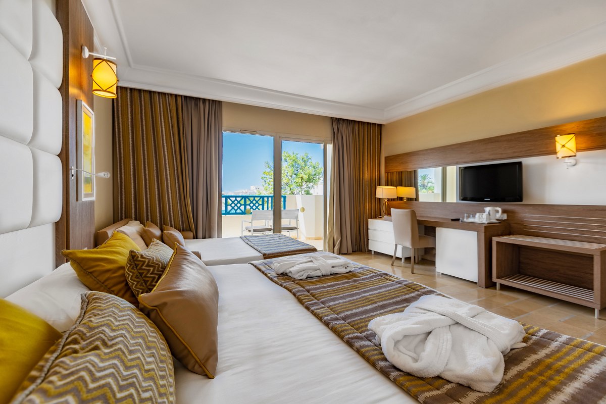 Modern hotel room with large bed, additional lounger, balcony with sea view and warm yellow-brown tones.