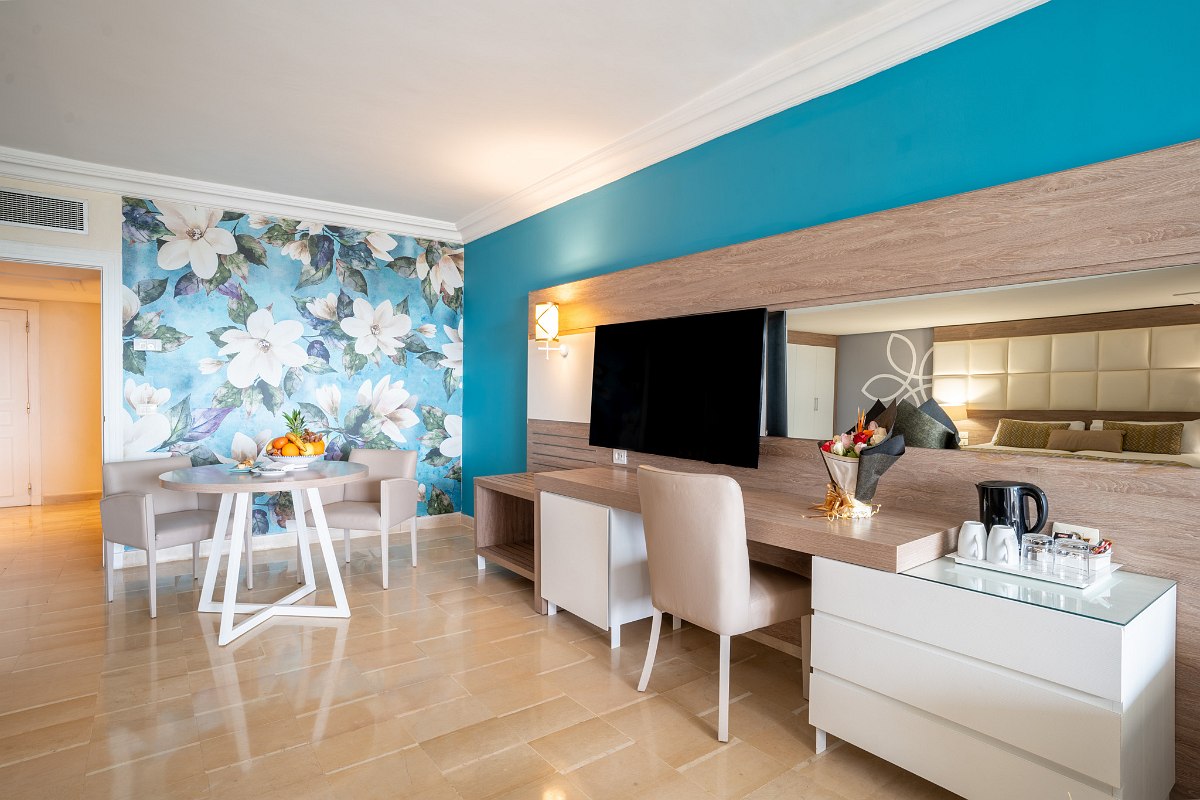 Bright hotel room with double bed, additional bed, balcony with sea view, desk and flat-screen TV.
