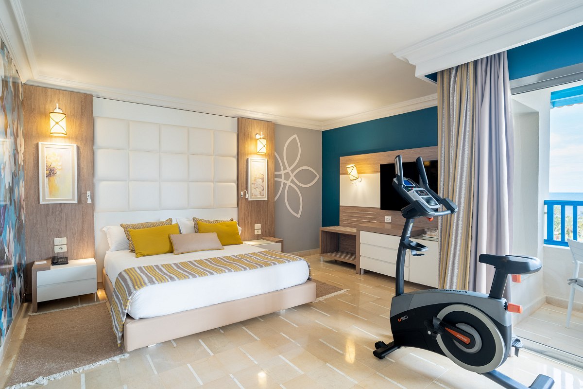 Spacious hotel room with bed, exercise bike, TV, balcony with sea view and fitness wall.