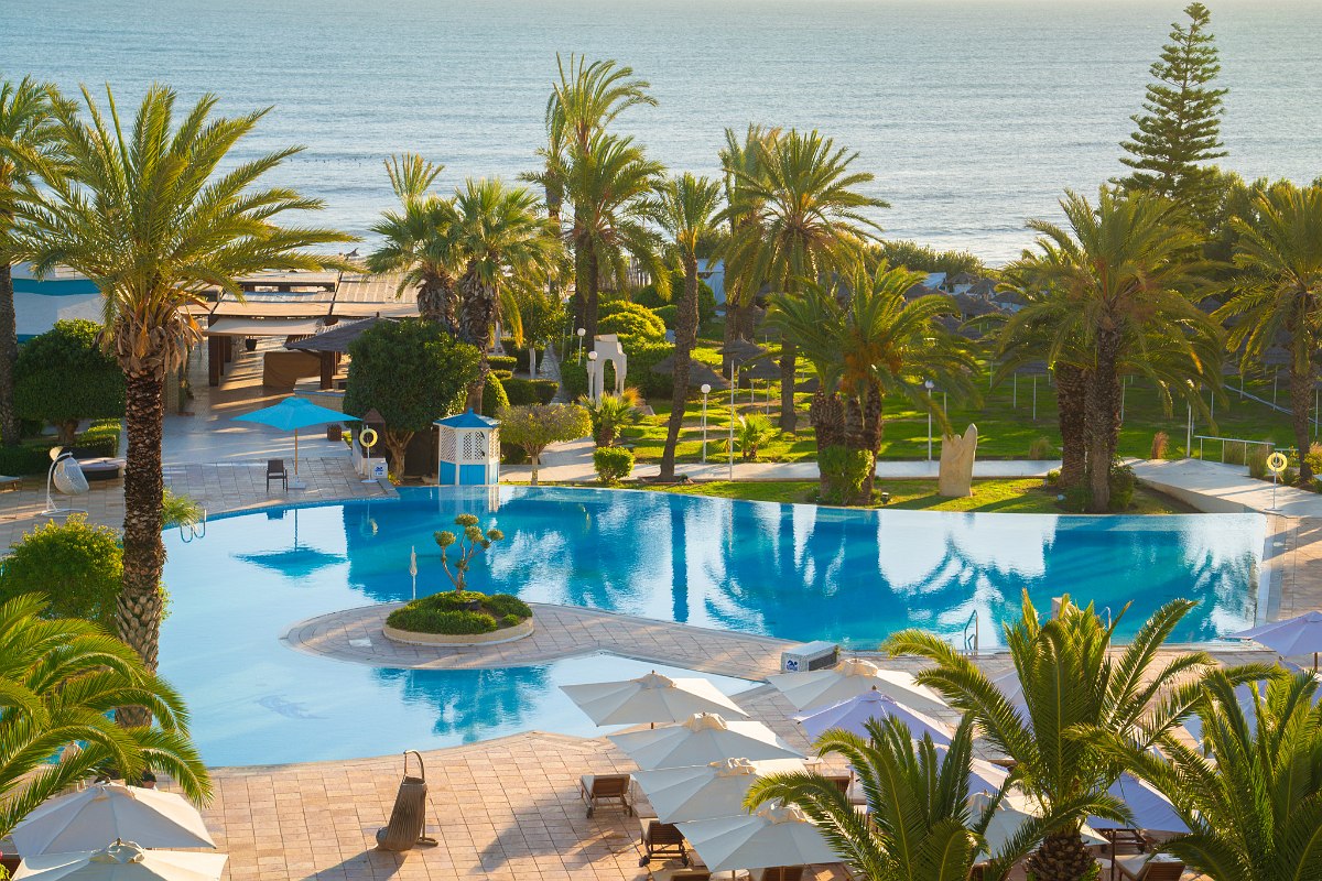 Large hotel pool with palm trees, sun loungers and a view of the Sentido Bellevue Park under a bright blue sky.