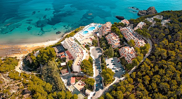Aerial view of a resort on a turquoise blue bay with beach, pool area and surrounded by dense pine forest.