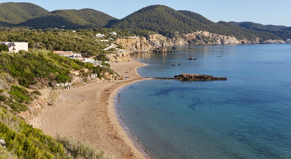 Quiet bay with a sandy beach and clear water, surrounded by green hills and rocky coastline in sunny weather.