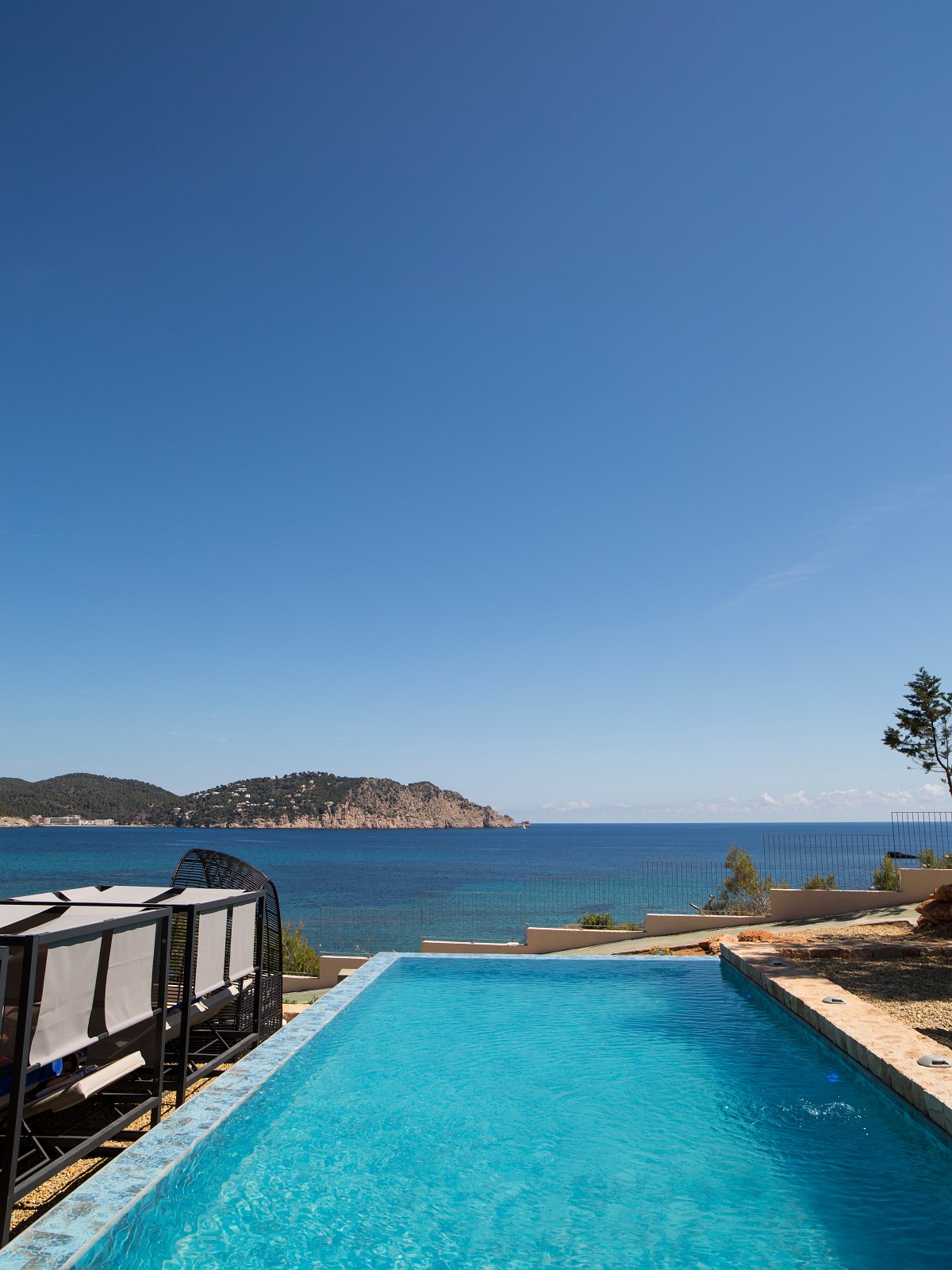 Infinity pool with sea view and sun loungers, green coastline and blue sky in the background.