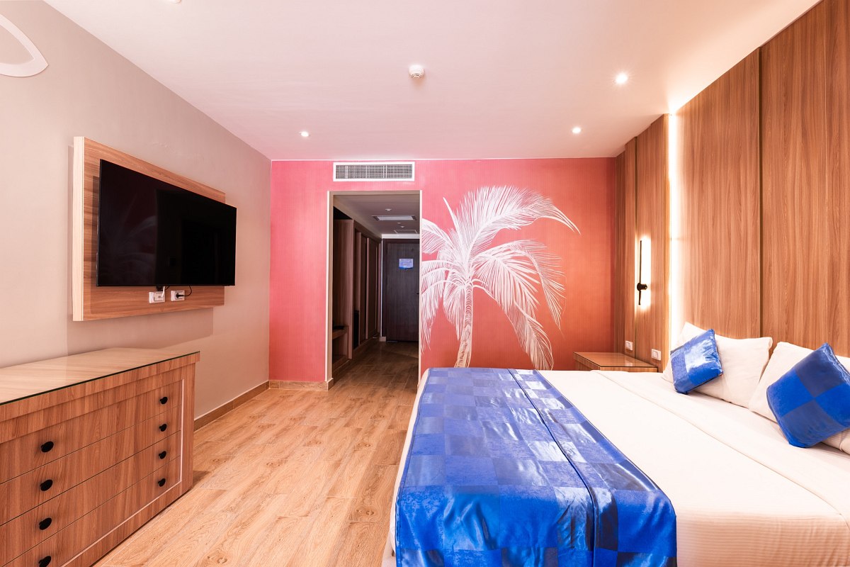 Modern room with a large bed, wood floors, blue accents, and a pink wall featuring a white palm tree design. Spacious and bright.
