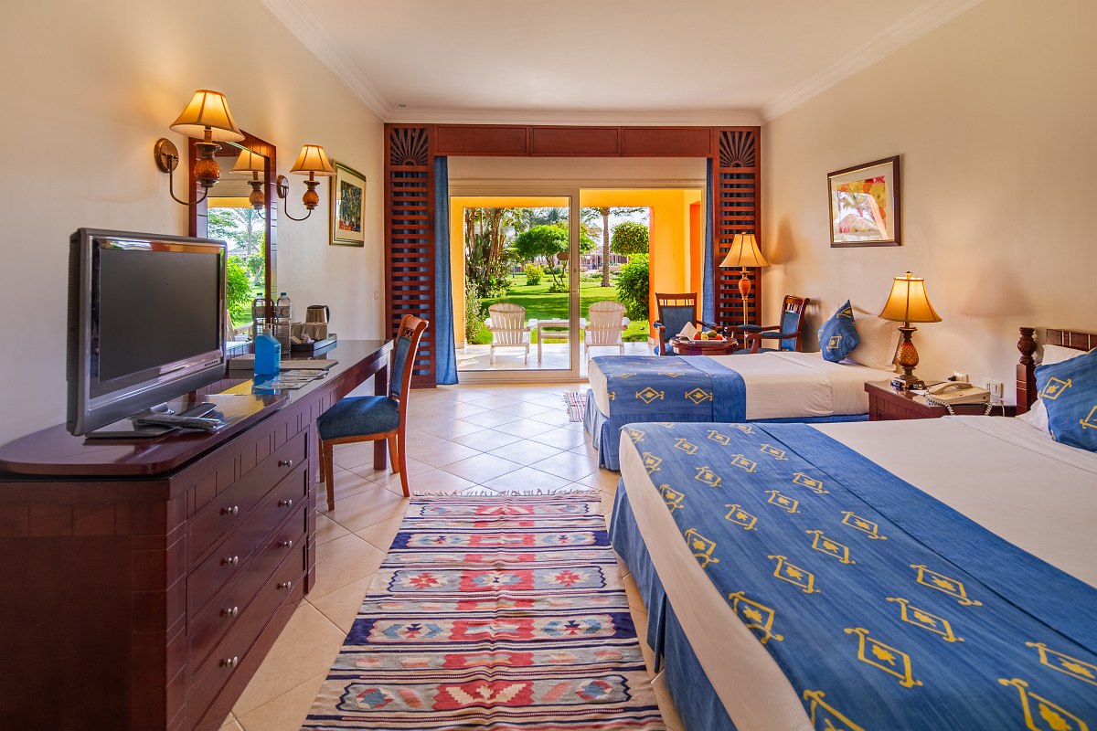 Cozy room with a large bed covered in blue bedding, a comfortable sofa, wooden desk and chair, and a balcony with outdoor seating and a view of greenery. Warm lighting adds to the inviting atmosphere.