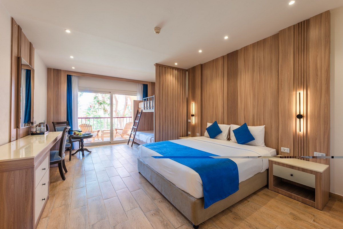 Modern room with wooden walls, large bed with blue accents, bunk beds, desk, TV, and balcony. Bright and spacious with warm lighting.